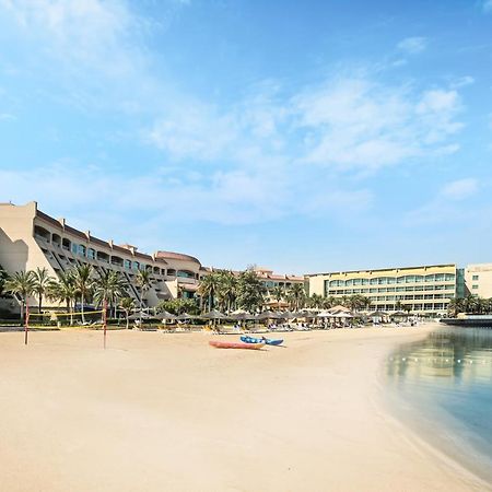 אבו דאבי Al Raha Beach Resort & Spa מראה חיצוני תמונה The photo depicts a serene beach scene with a clear blue sky. In the foreground, there is a sandy beach that gently slopes down to calm water. On the left side, several palm trees provide a tropical atmosphere. In the background, there are two large 