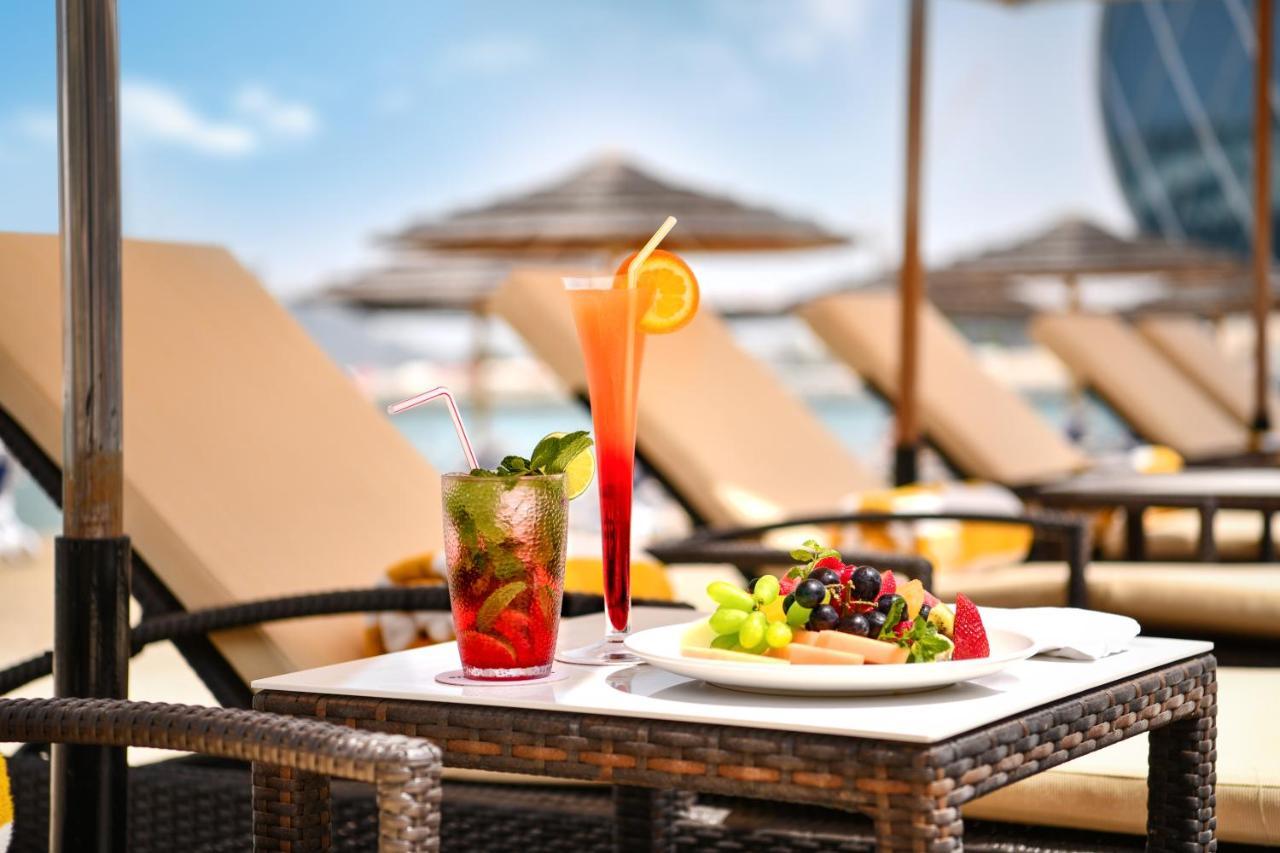 אבו דאבי Al Raha Beach Resort & Spa מראה חיצוני תמונה The photo shows a table set outdoors, featuring two colorful cocktails and a plate of food. One cocktail is a vibrant red drink garnished with mint, while the other is a layered orange beverage with an orange slice on the rim. The plate of food inclu
