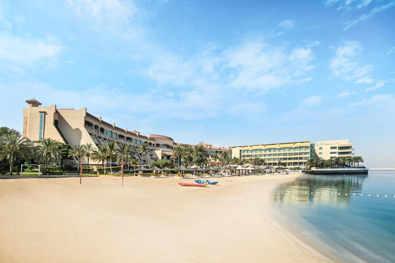 אבו דאבי Al Raha Beach Resort & Spa מראה חיצוני תמונה The photo depicts a serene beach scene with a clear blue sky. In the foreground, there is a sandy beach that gently slopes down to calm water. On the left side, several palm trees provide a tropical atmosphere. In the background, there are two large 