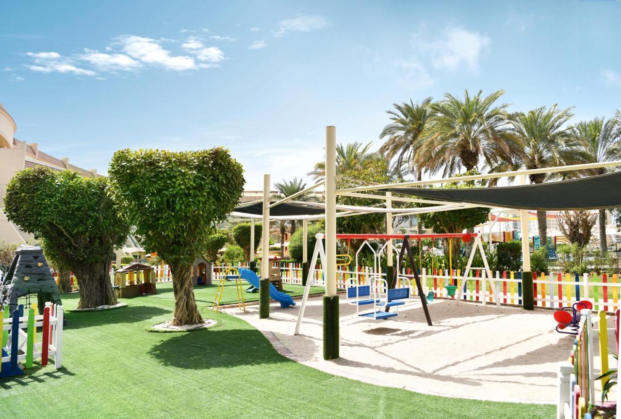 אבו דאבי Al Raha Beach Resort & Spa מראה חיצוני תמונה The photo shows a brightly lit outdoor playground surrounded by palm trees. The playground features a variety of play equipment, including swings, a slide, and a climbing structure. The ground is covered in lush green artificial grass, providing a so