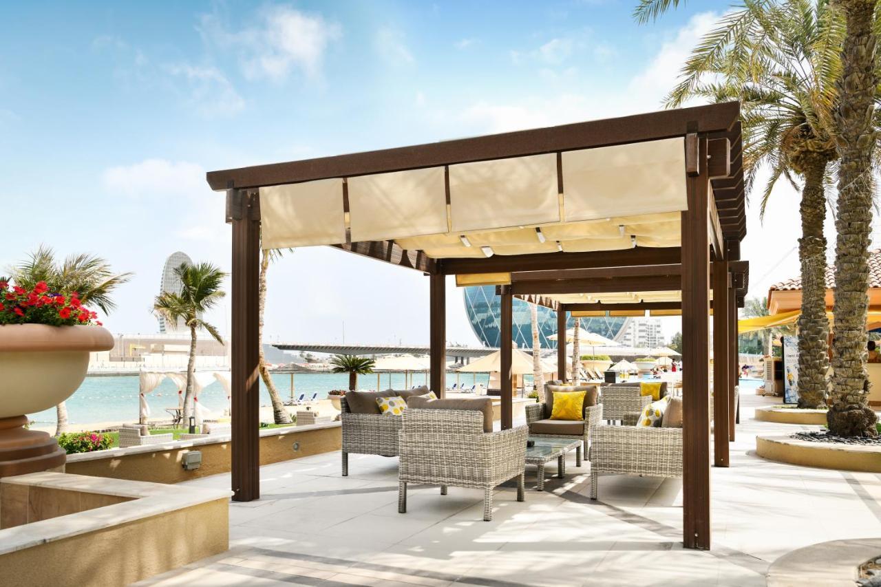 אבו דאבי Al Raha Beach Resort & Spa מראה חיצוני תמונה The photo shows a stylish outdoor seating area by a beach. There is a wooden pergola covered with a light fabric shade, providing shelter to a set of comfortable wicker chairs and tables. The area is adorned with palm trees, and in the background, th