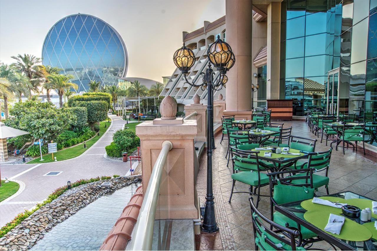 אבו דאבי Al Raha Beach Resort & Spa מראה חיצוני תמונה The photo depicts an outdoor dining area with green tables and chairs arranged on a patio. In the background, there is a large, distinctive building with a striking blue dome, surrounded by well-maintained landscaping, including trees and plants. The