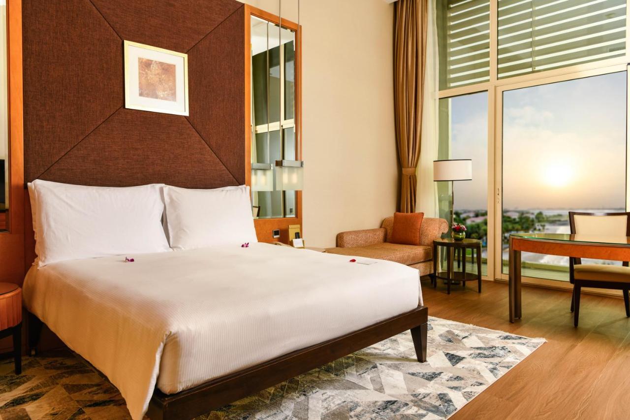אבו דאבי Al Raha Beach Resort & Spa מראה חיצוני תמונה The photo shows a stylish hotel room featuring a large bed with white bedding, positioned against a textured wall. There’s a small seating area with a chair and a table, and a window with curtains that allows natural light to fill the space. The room