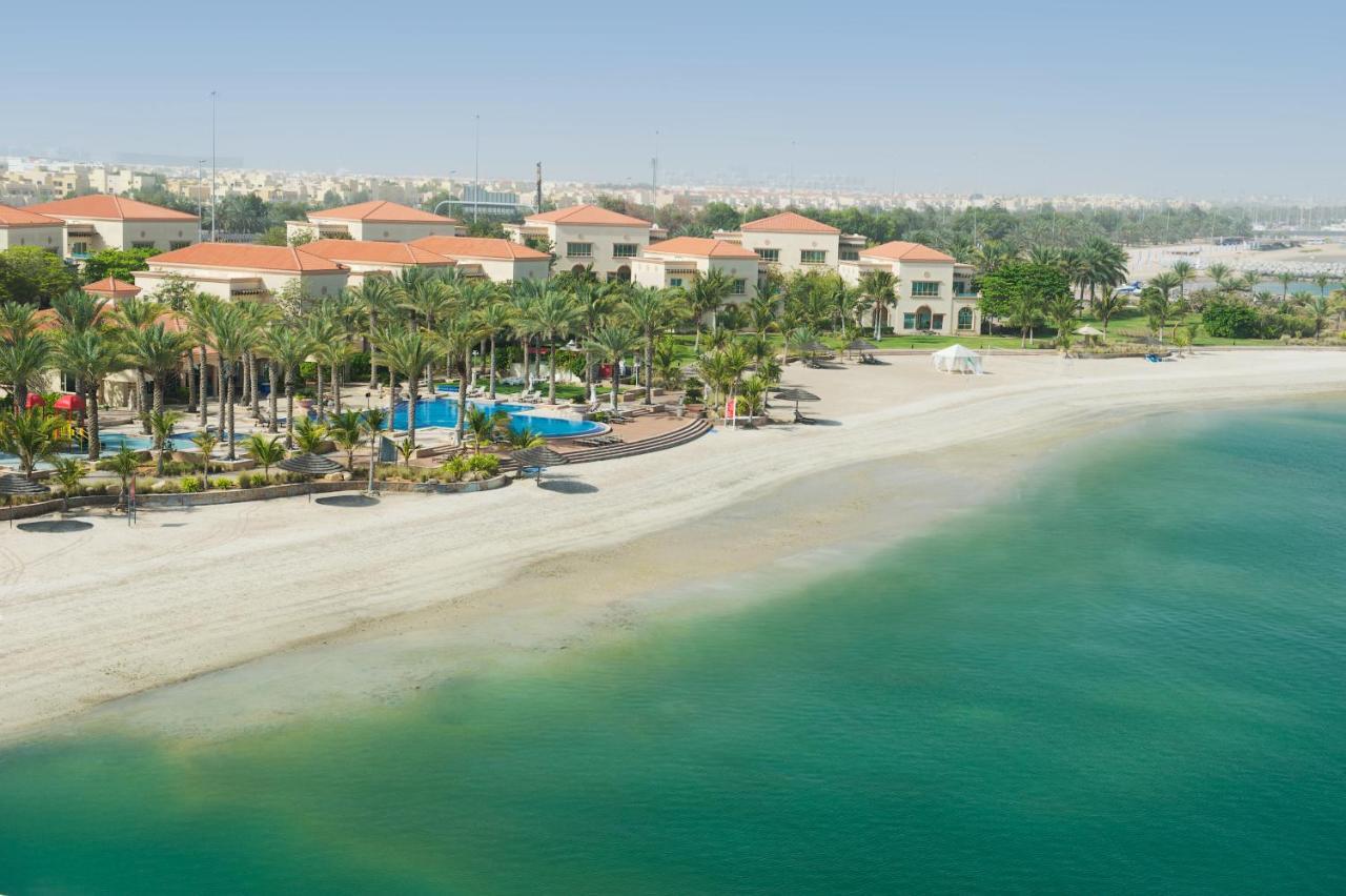 אבו דאבי Al Raha Beach Resort & Spa מראה חיצוני תמונה The photo depicts a serene beach scene with a clear turquoise sea and a sandy shoreline. In the background, there are several buildings featuring a Mediterranean architectural style with red-tiled roofs. The area is adorned with palm trees and outdoo