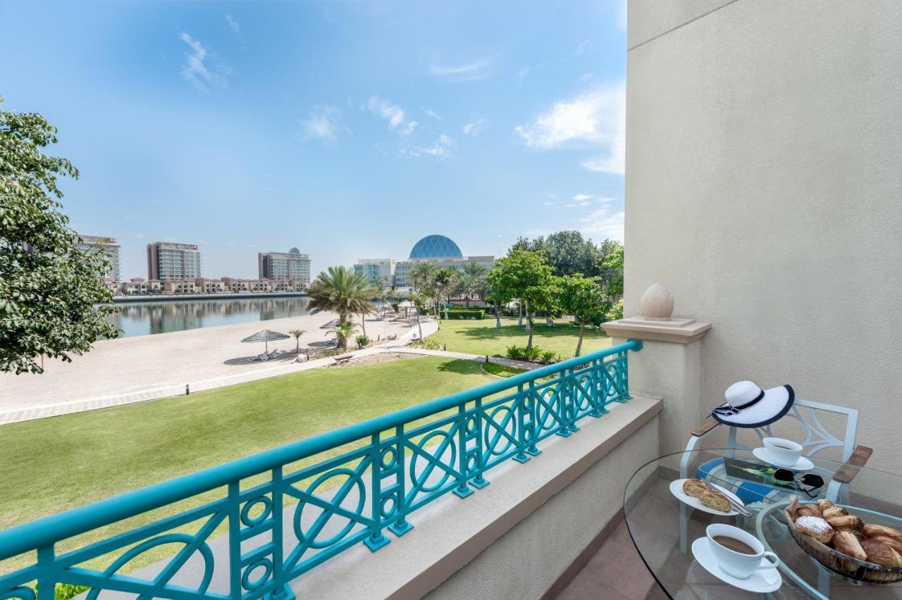 אבו דאבי Al Raha Beach Resort & Spa מראה חיצוני תמונה The photo shows a balcony view overlooking a scenic landscape. In the foreground, there is a glass table set with snacks or pastries and two cups. Beyond the balcony railing, there is a well-maintained green area with palm trees and a pathway. The ba