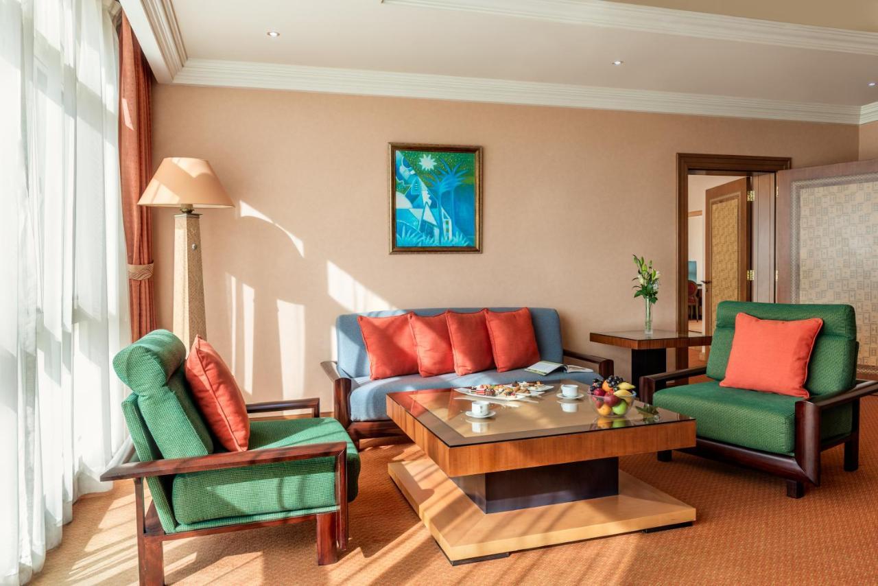 אבו דאבי Al Raha Beach Resort & Spa מראה חיצוני תמונה The photo shows a cozy living room with a warm ambiance. There are two sofas: one in green with red cushions and another in blue with red cushions, arranged around a wooden coffee table. On the table, there are some fruits and a small decorative item