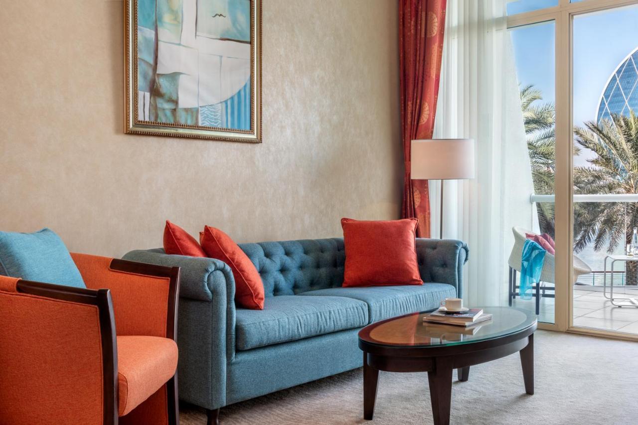 אבו דאבי Al Raha Beach Resort & Spa מראה חיצוני תמונה The photo shows a well-decorated living room. There is a plush blue couch with orange accent pillows and a cozy orange armchair nearby. A round coffee table sits in front of the couch, and above it, there is a colorful painting hanging on the wall. L