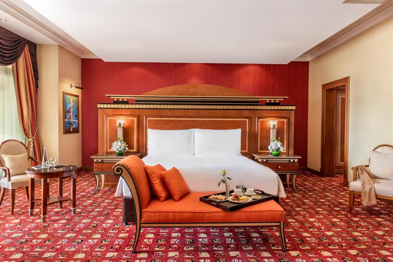 אבו דאבי Al Raha Beach Resort & Spa מראה חיצוני תמונה The photo shows an elegantly designed hotel room. The room features a large bed with white linens and a decorative headboard. The walls are painted in a warm red color, and there are side tables with lamps beside the bed. 

There is an orange sofa or
