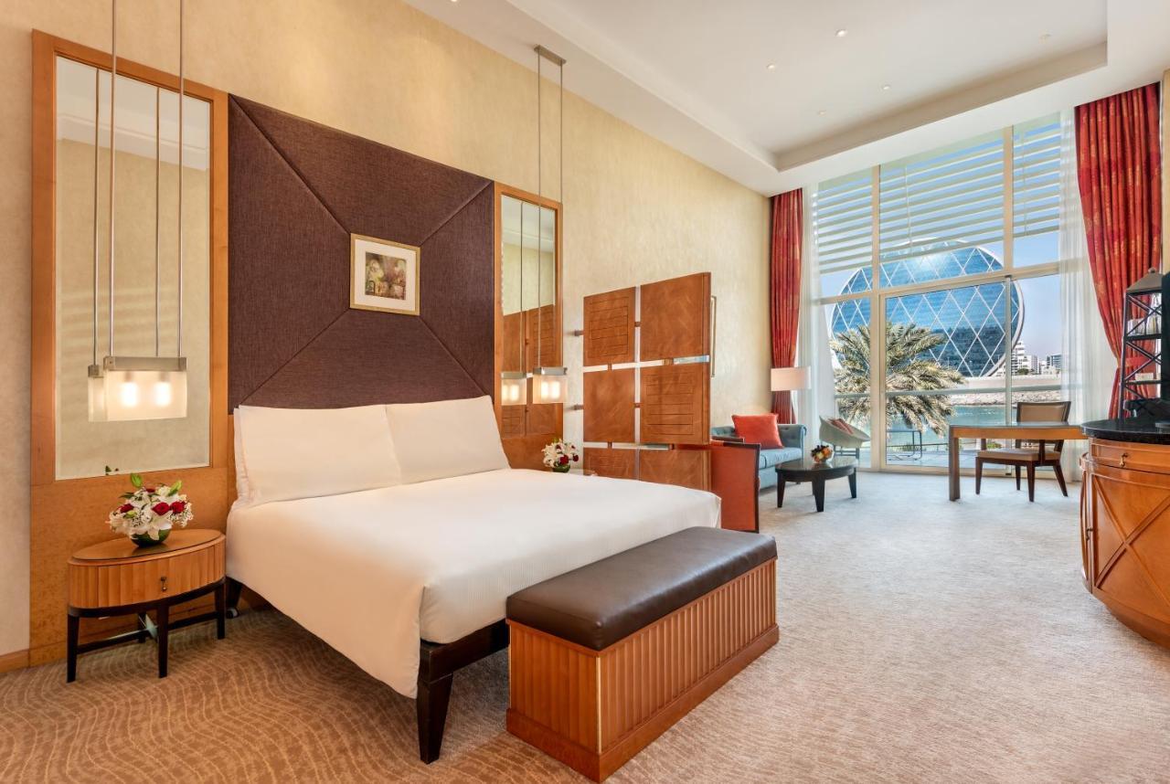 אבו דאבי Al Raha Beach Resort & Spa מראה חיצוני תמונה The photo shows a modern hotel room featuring a neatly made bed with a white comforter, situated against a stylish headboard with a geometric design. There are bedside tables on either side of the bed, each with a small flower arrangement. A comforta