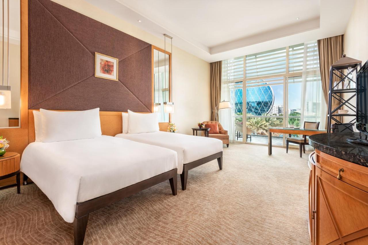 אבו דאבי Al Raha Beach Resort & Spa מראה חיצוני תמונה The photo shows a well-decorated hotel room featuring two single beds with white linens. The room has a modern aesthetic, with a large window letting in natural light and offering a view of a circular structure outside, possibly a landmark or artisti