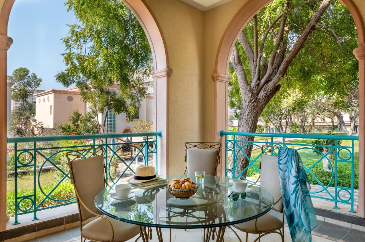 אבו דאבי Al Raha Beach Resort & Spa מראה חיצוני תמונה The photo shows a serene outdoor balcony setting. There is a circular glass table with a bowl of fruit or snacks in the center, alongside a glass and a small plate. Surrounding the table are four elegant chairs. The railing is turquoise, framing a vi