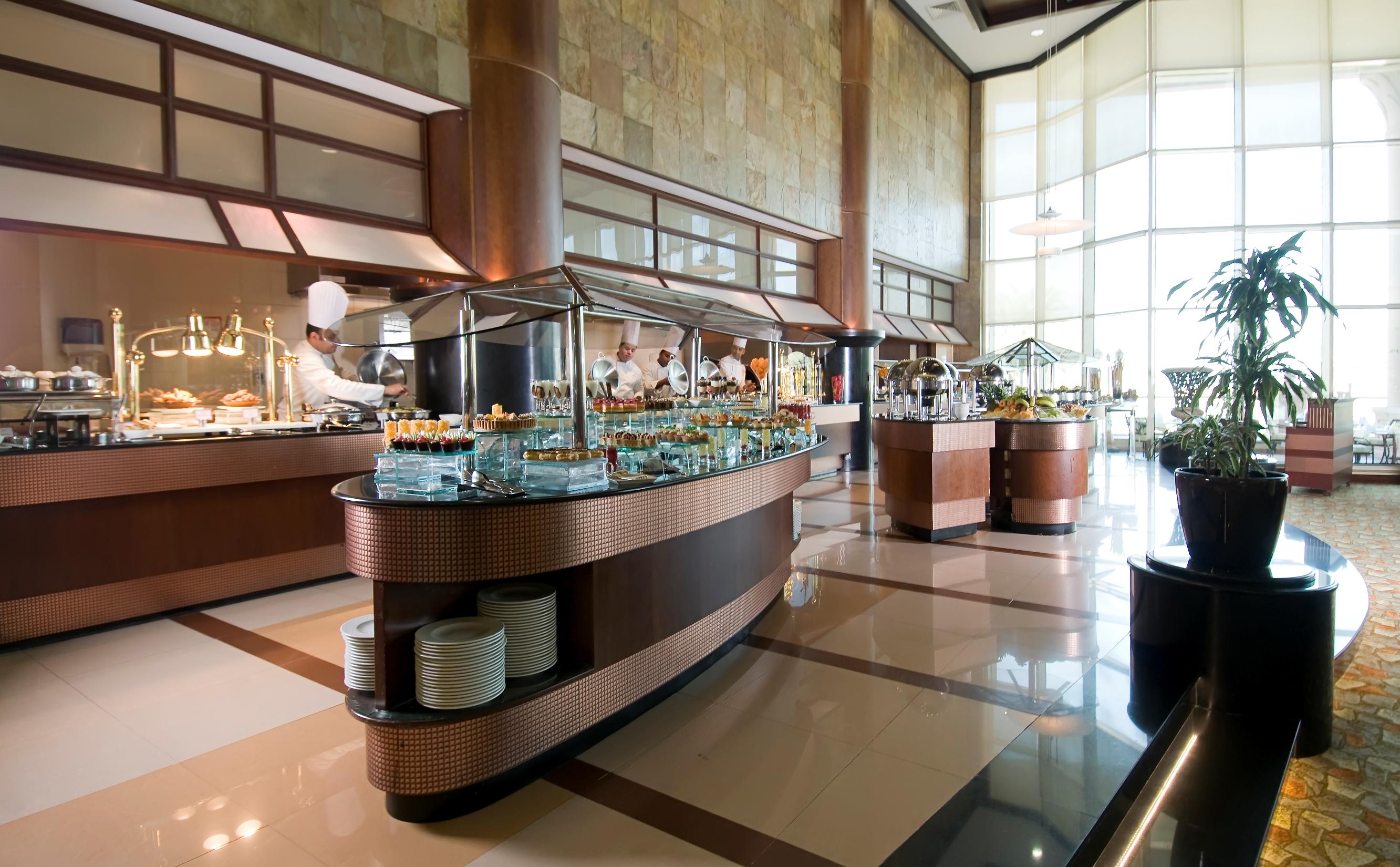 אבו דאבי Al Raha Beach Resort & Spa מראה חיצוני תמונה The photo shows a spacious buffet area in a restaurant or hotel. There are multiple food stations with glass displays showcasing a variety of dishes. Chefs can be seen in the background, preparing food. The decor features large windows allowing natur