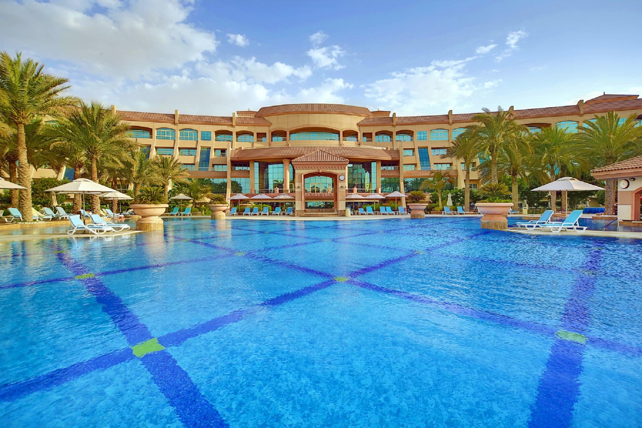 אבו דאבי Al Raha Beach Resort & Spa מתקנים תמונה The photo features a luxurious resort with a large swimming pool in the foreground. The pool is crystal clear and reflects the sky, bordered by lounge chairs and palm trees. In the background, the resort building showcases modern architecture with la