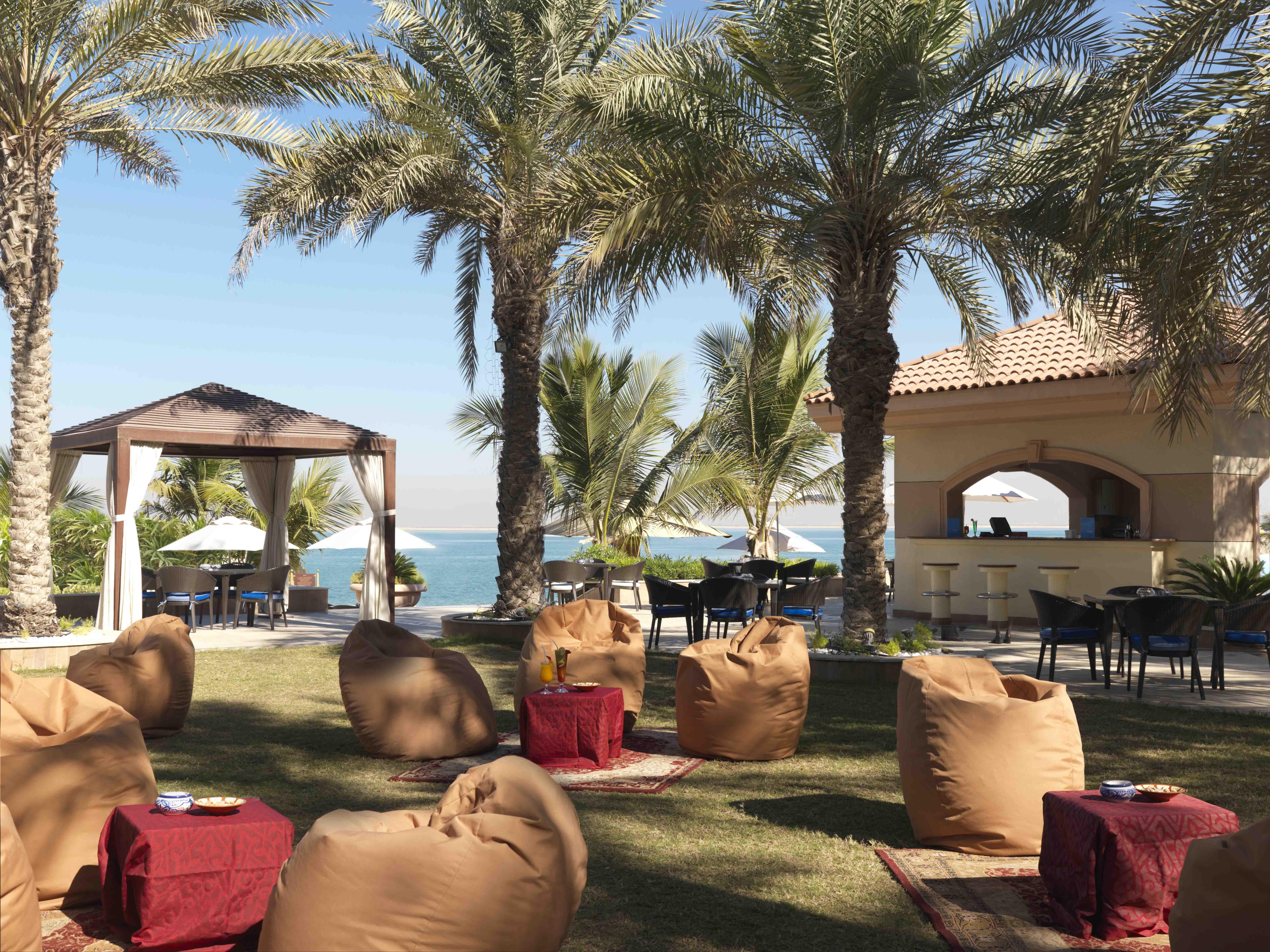 אבו דאבי Al Raha Beach Resort & Spa מסעדה תמונה The photo depicts a beautiful outdoor setting with several elements of relaxation and leisure. In the foreground, there are large, comfortable bean bag chairs arranged on a grassy area, accompanied by low tables. Surrounding this seating area are tal