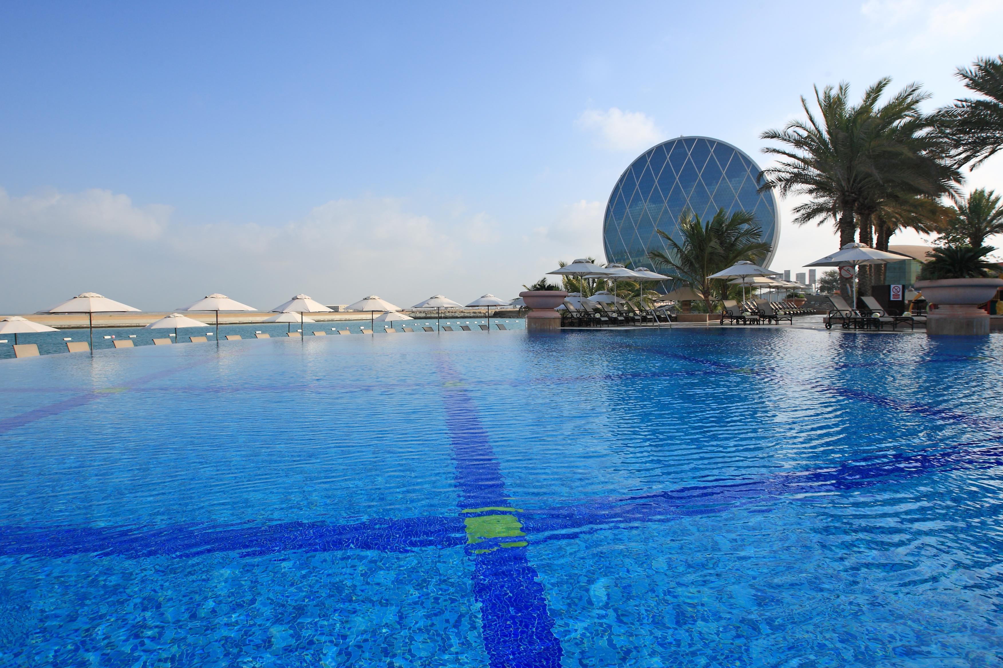 אבו דאבי Al Raha Beach Resort & Spa מראה חיצוני תמונה The photo features a large, serene swimming pool with clear, blue water reflecting the sky. Surrounding the pool are several lounge chairs and umbrellas, suggesting a relaxed, resort-like atmosphere. In the background, a distinctive building with a r