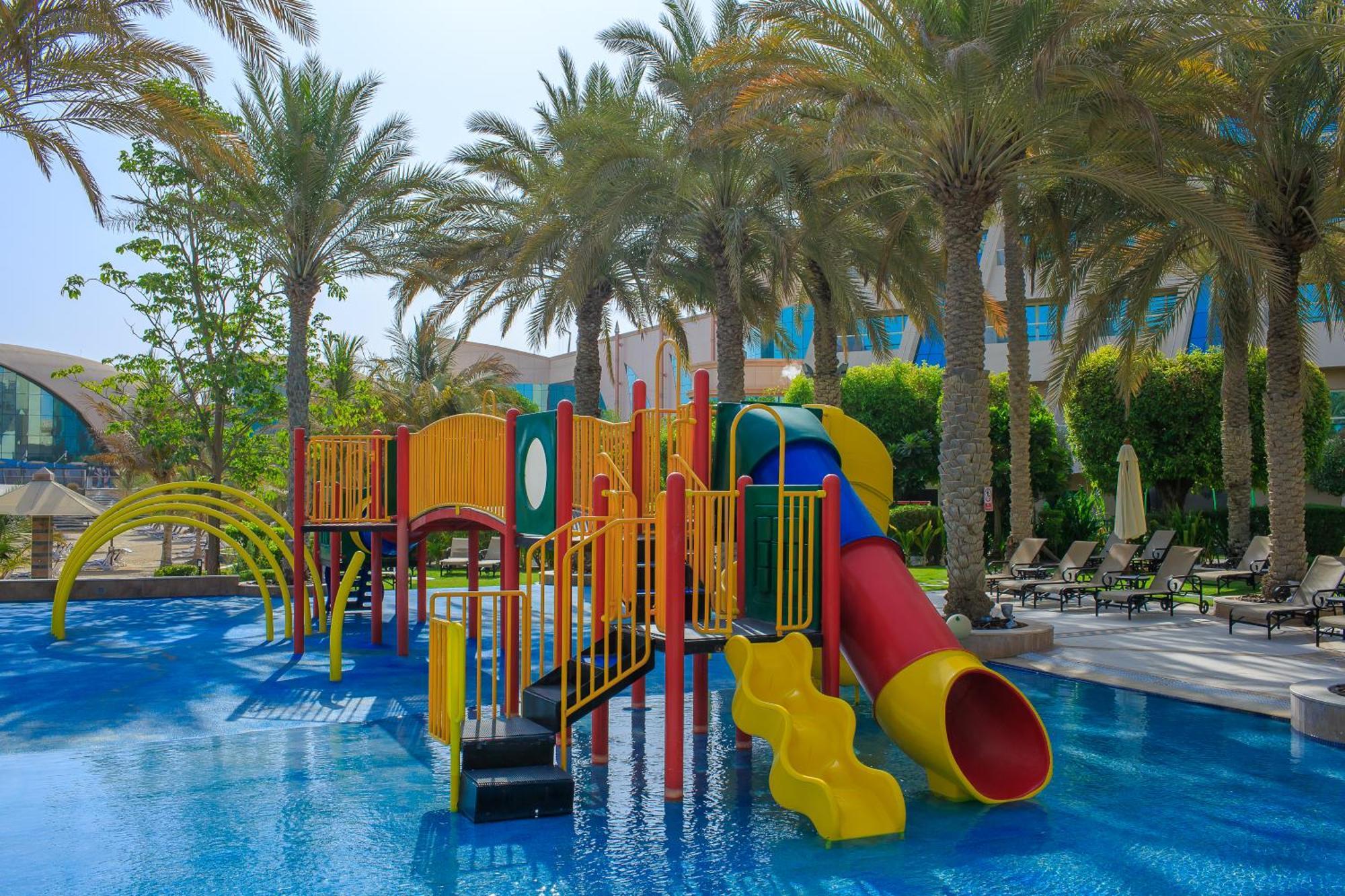 אבו דאבי Al Raha Beach Resort & Spa מראה חיצוני תמונה The photo shows a colorful water play area located in a pool setting. It features a variety of play structures, including slides in multiple colors, such as yellow, red, and blue. Surrounding the play structure are palm trees, creating a tropical atm