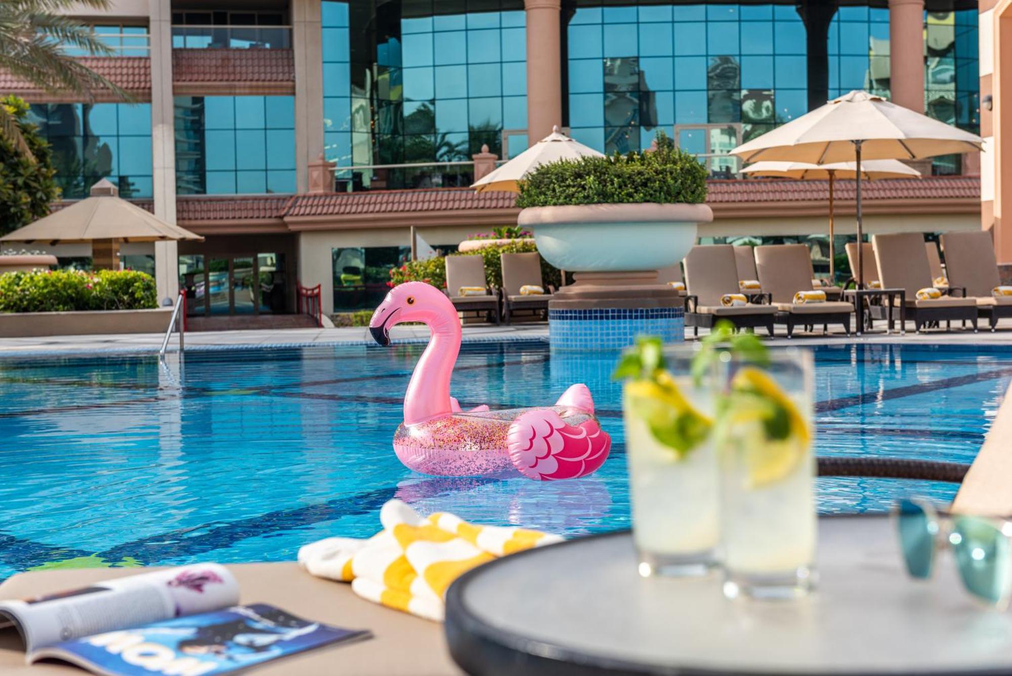 אבו דאבי Al Raha Beach Resort & Spa מראה חיצוני תמונה The photo depicts a bright and inviting outdoor swimming pool surrounded by a luxurious setting. In the pool, there is a large pink flamingo float, adding a fun and whimsical touch. To the side, a table is set up with two refreshing drinks, likely co
