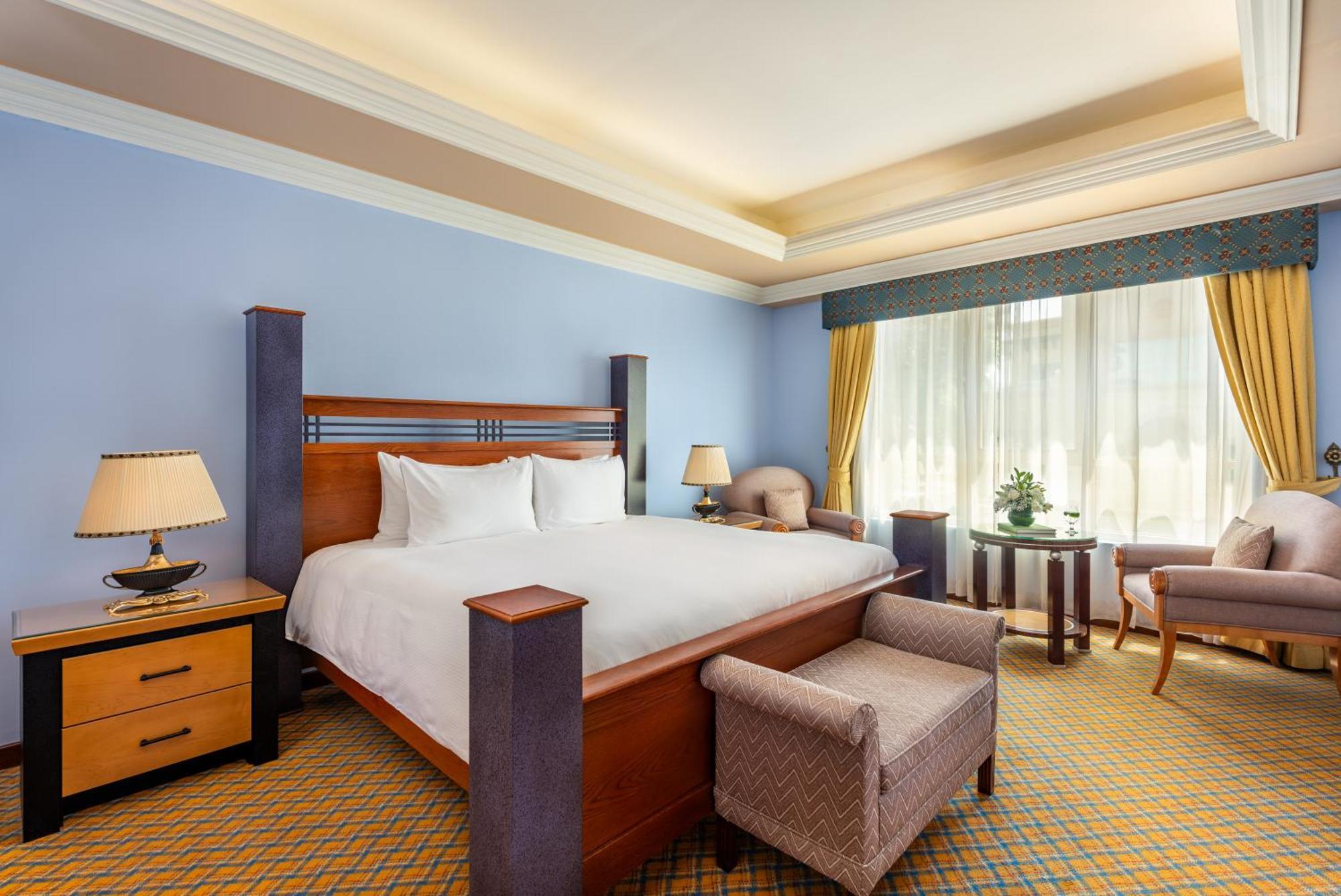 אבו דאבי Al Raha Beach Resort & Spa מראה חיצוני תמונה The photo shows a well-decorated hotel room. It features a large bed with a wooden frame, neatly made with white bedding. On either side of the bed are bedside tables with lamps. There is a comfortable chair next to a window that has sheer curtains a