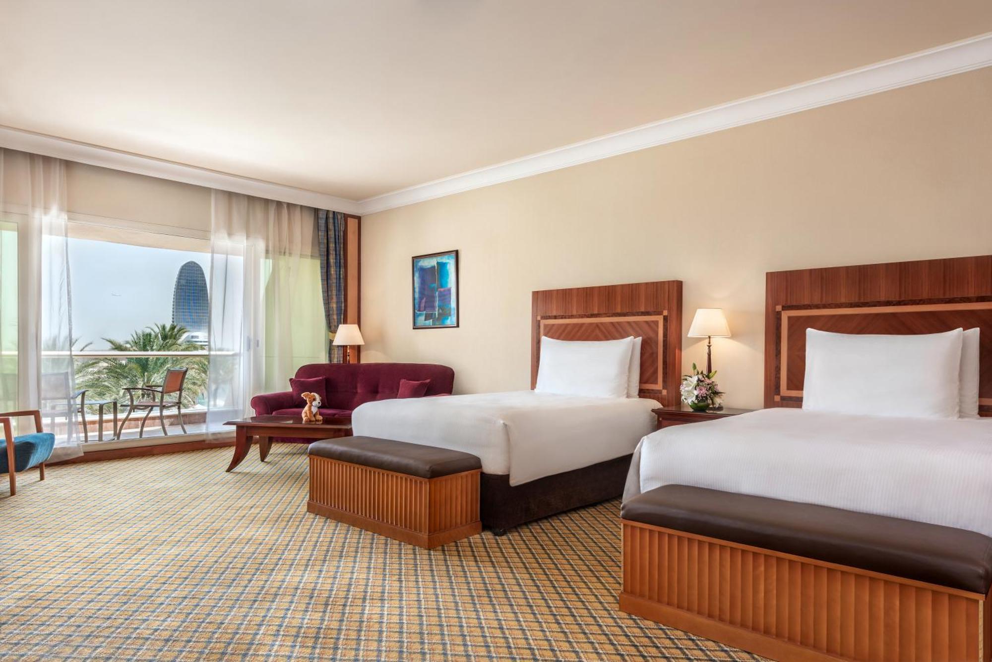 אבו דאבי Al Raha Beach Resort & Spa חדר תמונה The photo shows a hotel room featuring two twin beds with wooden headboards. The room has a light, neutral color scheme, with a large window that provides natural light and a view of the outdoors. There is a small seating area with a purple sofa and 