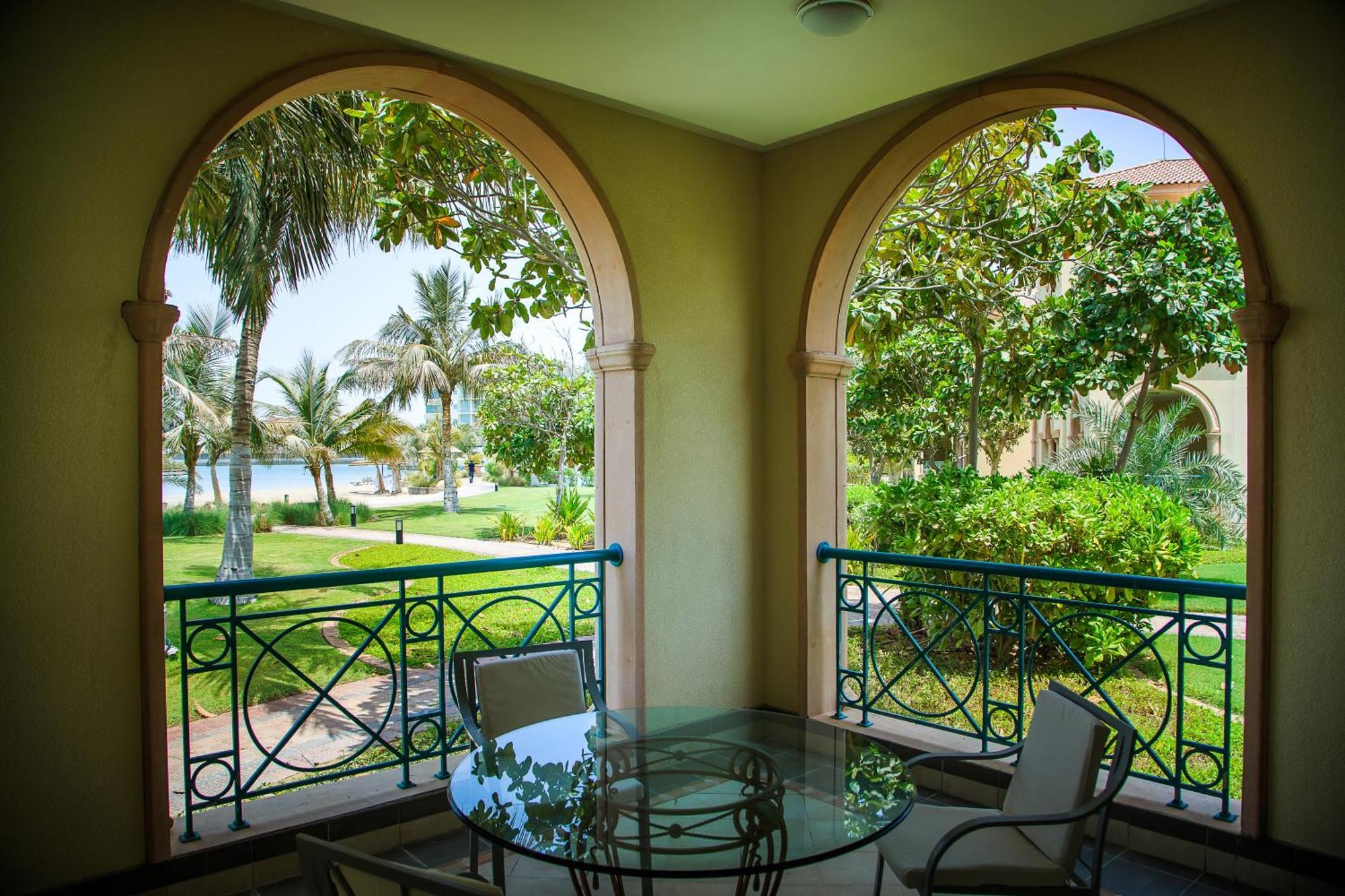אבו דאבי Al Raha Beach Resort & Spa חדר תמונה The photo displays a beautifully designed balcony or terrace area. It features two large arches that frame the view outside. Through the arches, you can see a lush garden with palm trees and well-maintained greenery. There’s a glass table with severa