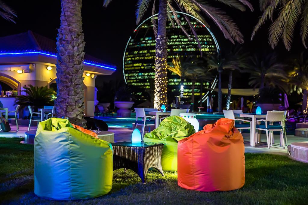אבו דאבי Al Raha Beach Resort & Spa מראה חיצוני תמונה The photo shows a vibrant outdoor lounge area at night. In the foreground, there are colorful bean bag chairs—one in bright green, one in yellow, and one in orange. A small table sits between the bean bags. In the background, illuminated palm trees a