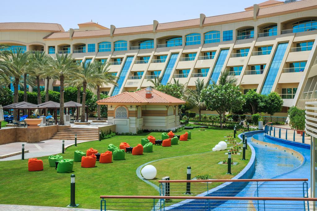 אבו דאבי Al Raha Beach Resort & Spa מראה חיצוני תמונה The photo depicts a modern hotel complex with multiple stories and large windows. In the foreground, there is a well-maintained lawn featuring colorful lounge chairs in shades of orange and green. A pathway runs alongside a blue water feature, creati