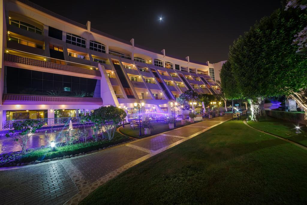 אבו דאבי Al Raha Beach Resort & Spa מראה חיצוני תמונה The photo shows a modern building illuminated at night, with a vibrant purple hue. The structure has multiple floors, each with balconies overlooking a landscaped area. In the foreground, there is a pathway lined with well-manicured grass and shrubs,