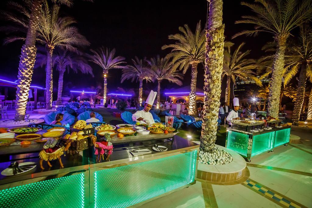 אבו דאבי Al Raha Beach Resort & Spa מראה חיצוני תמונה The photo depicts an outdoor dining area at night, featuring several chefs in white uniforms and hats working at a food station. The setting is illuminated by colorful lights, with glowing green accents on the serving tables. Surrounding the area are